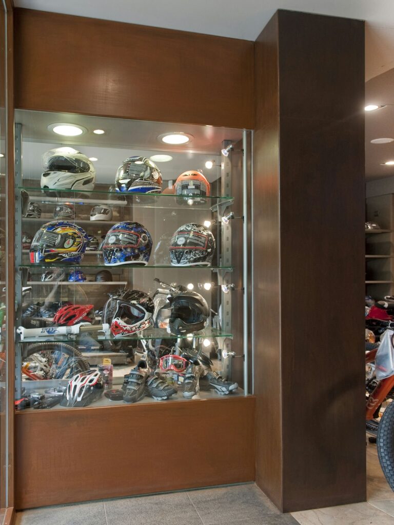 Exposición de Casco de Motocicleta y Equipamiento Deportivo en Diseño Moderno Vitrina contemporánea que muestra una variedad de cascos de motocicleta y equipamiento deportivo, iluminada con suaves luces y con estantes de vidrio en un entorno de madera elegante.
