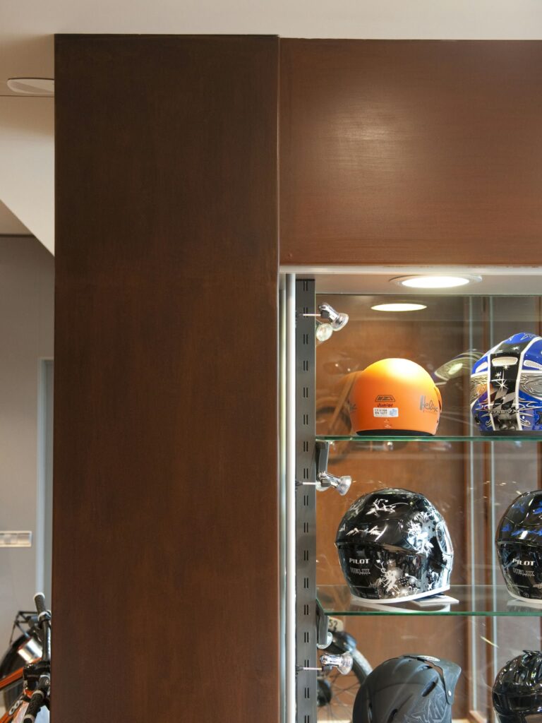 Exhibición de Casquitos Diversos en un Vitrina Moderna Vitrina iluminada que muestra varios cascos, incluyendo un casco naranja vibrante y varios cascos negros diseñados de manera única. La vitrina está hecha de vidrio y metal, situada contra un fondo contemporáneo con un panel de madera a la izquierda.