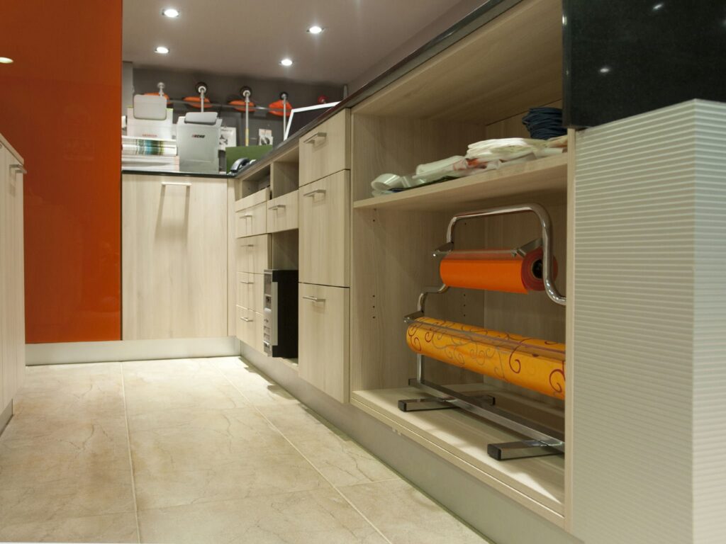Interior de Cocina Moderna con Muebles Elegantes y Acentos Vibrantes Un interior de cocina moderna que muestra armarios de madera de colores claros con un diseño elegante, una pared de acento naranja vibrante y un suelo de baldosas limpio, con soluciones de almacenamiento organizadas para utensilios y un rollo naranja en un soporte de metal.