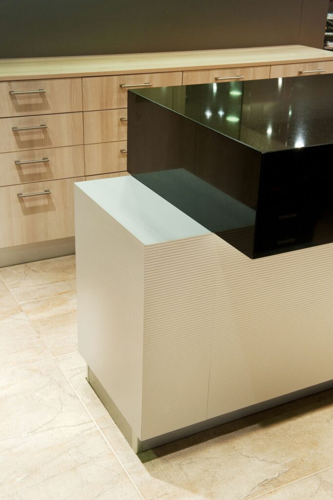 Diseño de Cocina Moderna y Elegante con Gabinetes de Madera Clara y Encimera Negra Cocina moderna que muestra gabinetes de madera clara con manijas metálicas y una llamativa encimera negra sobre una base blanca texturizada. El espacio presenta pisos de cerámica de color claro y un fondo minimalista, destacando líneas limpias y materiales contemporáneos para una estética elegante.