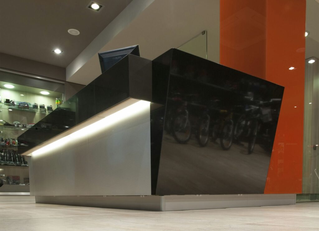 Recepción moderna con diseño elegante en negro y acentos naranjas Un mostrador de recepción contemporáneo hecho de material negro brillante con formas angulares y una base iluminada, situado frente a un fondo de acentos naranjas brillantes y estantes que exhiben varios dispositivos electrónicos. La suave iluminación superior crea una atmósfera acogedora, mientras que un monitor de computadora en el mostrador indica su uso para el servicio al cliente.