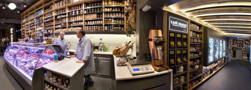 Diseño de Interiores Moderno en Barcelona: Espacio de Especialidad de Alta Gama Interior de una tienda moderna y de alta gama en Barcelona, exhibiendo un diseño funcional y estético con mostrador elegante, productos frescos en una vitrina de cristal, estanterías de madera con tarros gourmet, y mobiliario personalizado en iluminación natural.