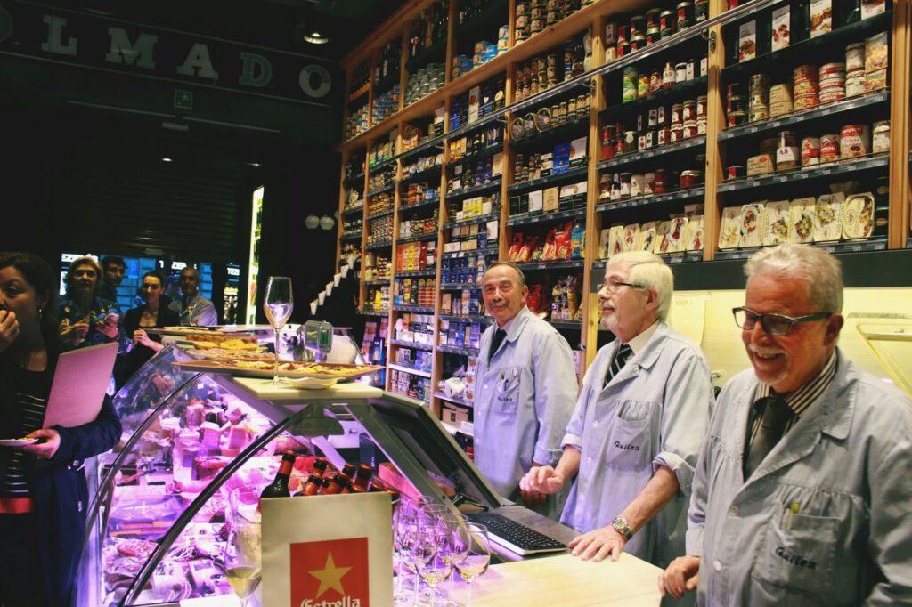Mercado Tradicional en Barcelona: Cultura Gastronómica Local Vendedores en un mercado de comida en Barcelona, exhibiendo productos gourmet en un entorno vibrante con estantes llenos de productos variados.