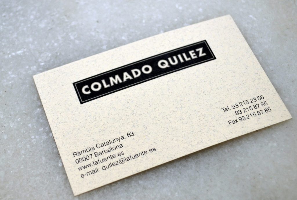 Tarjeta de visita moderna de Colmado Quilez en Barcelona Primer plano de la tarjeta de visita de Colmado Quilez, destacando un diseño limpio y moderno en un entorno de superficie texturizada. La tarjeta incluye dirección, números de teléfono y correo electrónico, reflejando la estética y funcionalidad del diseño de interiores y arquitectura de alta gama en Barcelona.