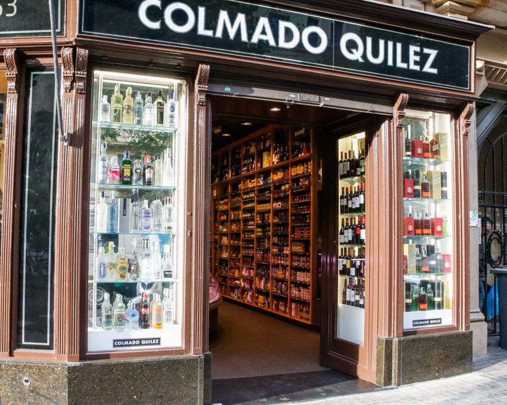 Colmado Quilez: Tienda de lujo en Barcelona con elegancia arquitectónica Fachada del Colmado Quilez en Barcelona, destacando sus vitrinas de vidrio con botellas de licores y vinos en un entorno sofisticado y moderno.