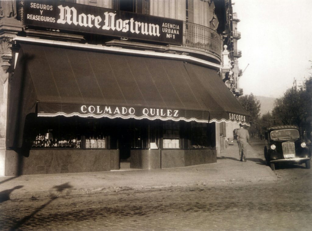 Escena Histórica en Barcelona: Colmado Quliez y su Herencia Cultural Escena histórica de la calle en Barcelona que muestra la tienda 'Colmado Quliez' con un toldo elegante, un coche vintage estacionado y un transeúnte, resaltando la arquitectura de la ciudad.