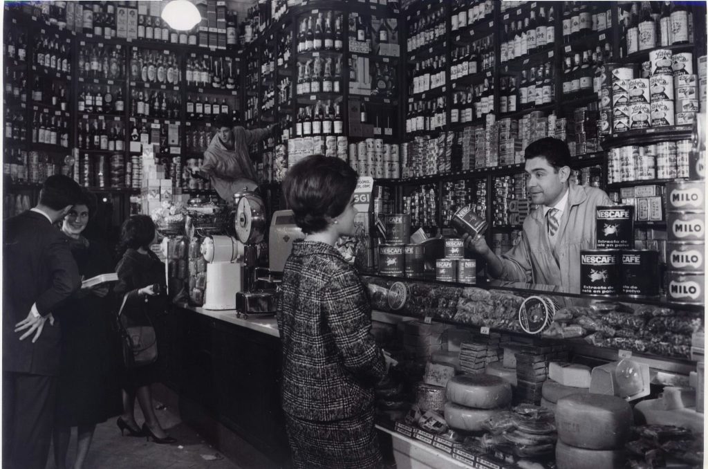Interiores de una tienda de comestibles vintage del siglo XX Interior de una tienda de comestibles vintage con estanterías llenas de productos, un shopkeeper atendiendo a una cliente, y un ambiente nostálgico en fotografía en blanco y negro.