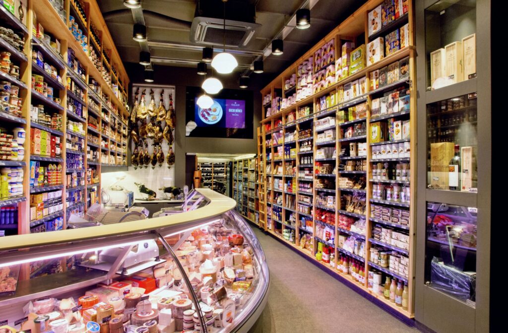 Proyecto de Diseño de Interiores en Barcelona - Gourmet Food Store Estanterías de madera organizadas en un gourmet food store en Barcelona, mostrando productos como latas, especias y embutidos, con una iluminación moderna y un mostrador curvo con quesos.