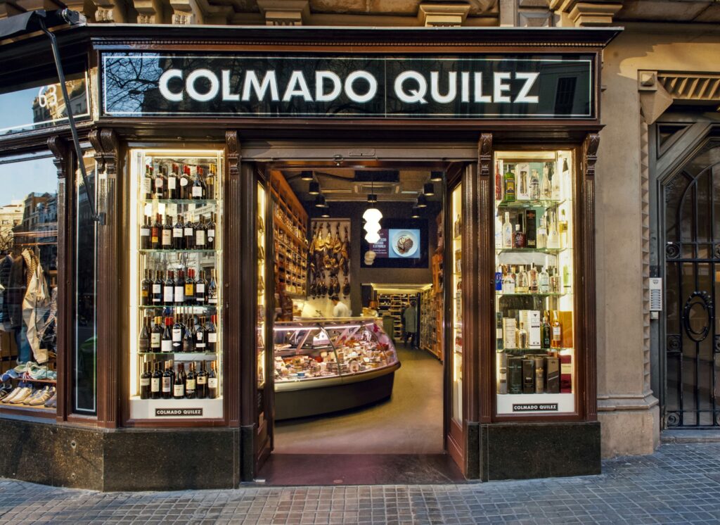 Colmado Quilez: Exterior de una tienda gourmet en Barcelona Vista exterior de Colmado Quilez en Barcelona, mostrando un moderno escaparate de supermercado gourmet con una variedad de vinos a la izquierda y productos de delicatessen a la derecha. El diseño resalta materiales elegantes, iluminación sofisticada y una atmósfera acogedora, reflejando la arquitectura y el diseño de interiores de alta gama.