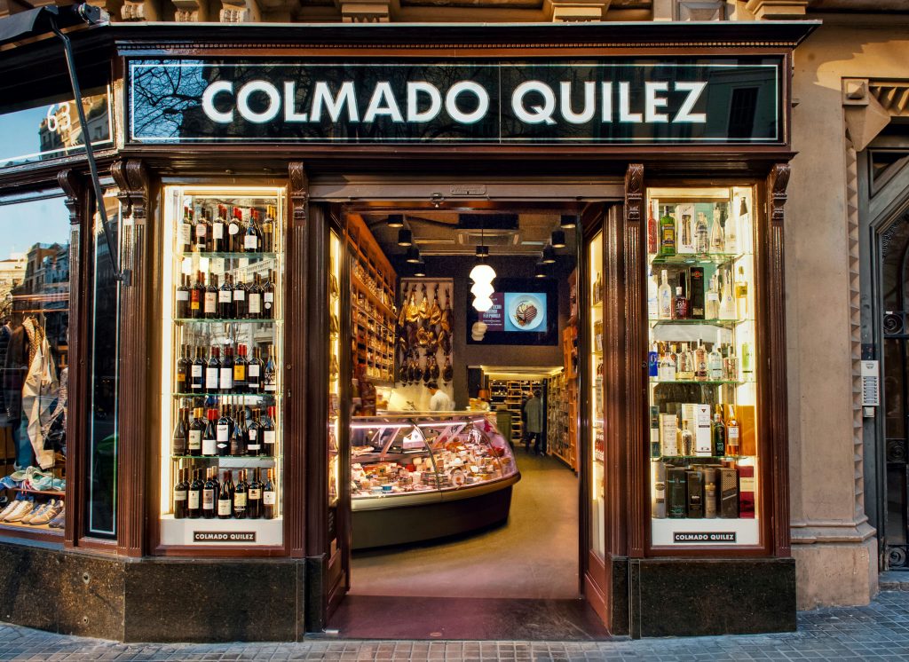 Vista exterior de COLMADO QUILEZ, una delicatessen de alta gama en Barcelona, con marcos de madera elegantes y grandes vitrinas de cristal exhibiendo una variedad de vinos y productos gourmet.