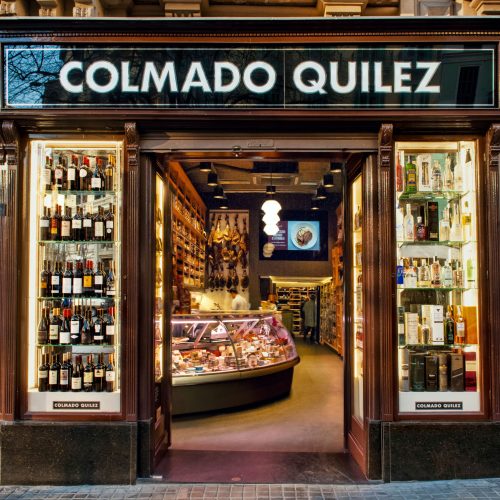 Vista exterior de COLMADO QUILEZ, una delicatessen de alta gama en Barcelona, con marcos de madera elegantes y grandes vitrinas de cristal exhibiendo una variedad de vinos y productos gourmet.