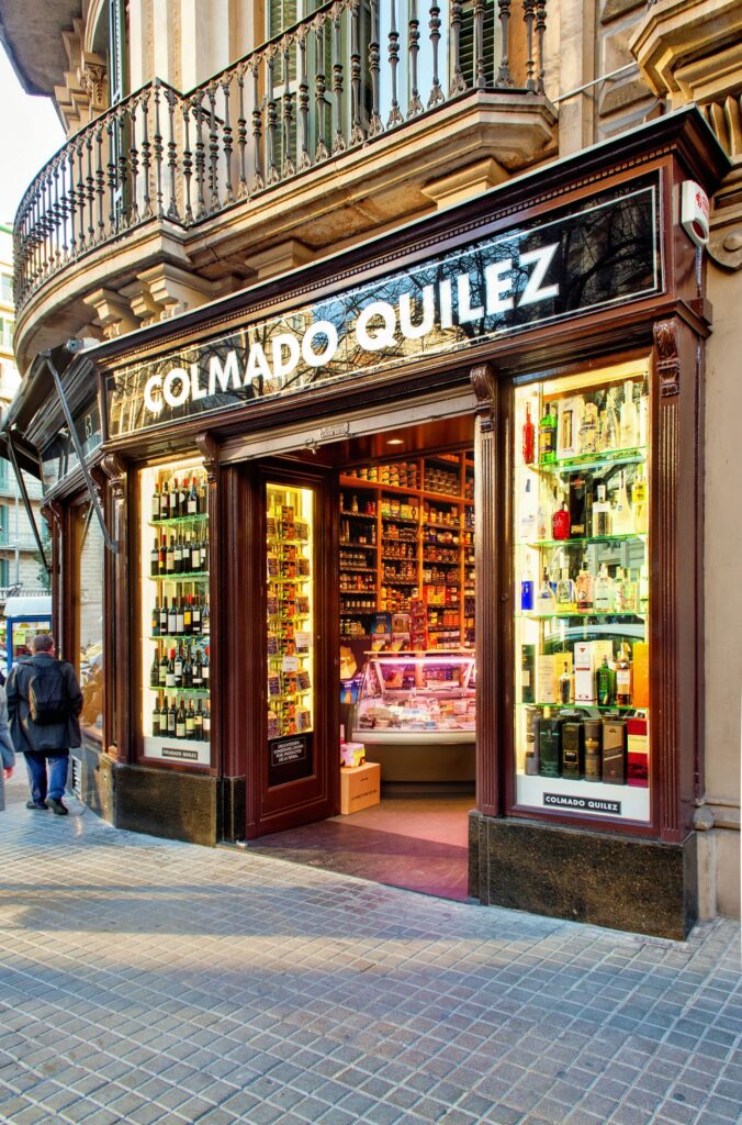 Colmado Quilez: Tienda Gourmet de Diseño Moderno en Barcelona Vista exterior de Colmado Quilez, una tienda de comestibles de alta gama en Barcelona con diseño sofisticado, grandes ventanales exhibiendo vinos y productos gourmet, en una calle empedrada.