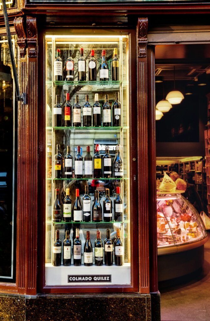 Vitrina de Botellas de Vino en Tienda de Barcelona con Diseño Moderno Vitrina iluminada en una tienda de Barcelona que exhibe una variedad de botellas de vino de alta gama en estantes elegantes, resaltando el diseño interior contemporáneo y acogedor.