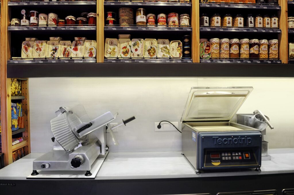 Cocina Moderna con Cortadora de Carne y Sellador de Vacío Tecnorip en Barcelona Contador de cocina moderno con cortadora de carne gris y sellador de vacío Tecnorip, respaldado por estantería de madera con tarros gourmet, reflejando diseño contemporáneo y funcionalidad.