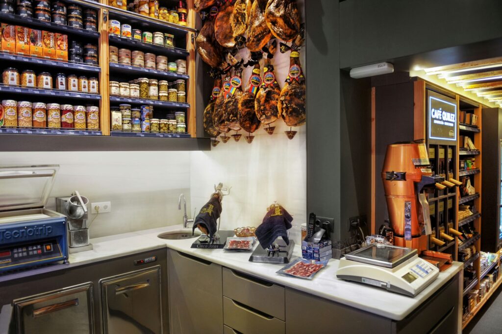 Interior de una tienda gourmet en Barcelona: Diseño y Funcionalidad Interior moderno de un delicatessen en Barcelona con estanterías de madera, productos gourmet y jamones españoles colgados, resaltando la calidad en el diseño arquitectónico.