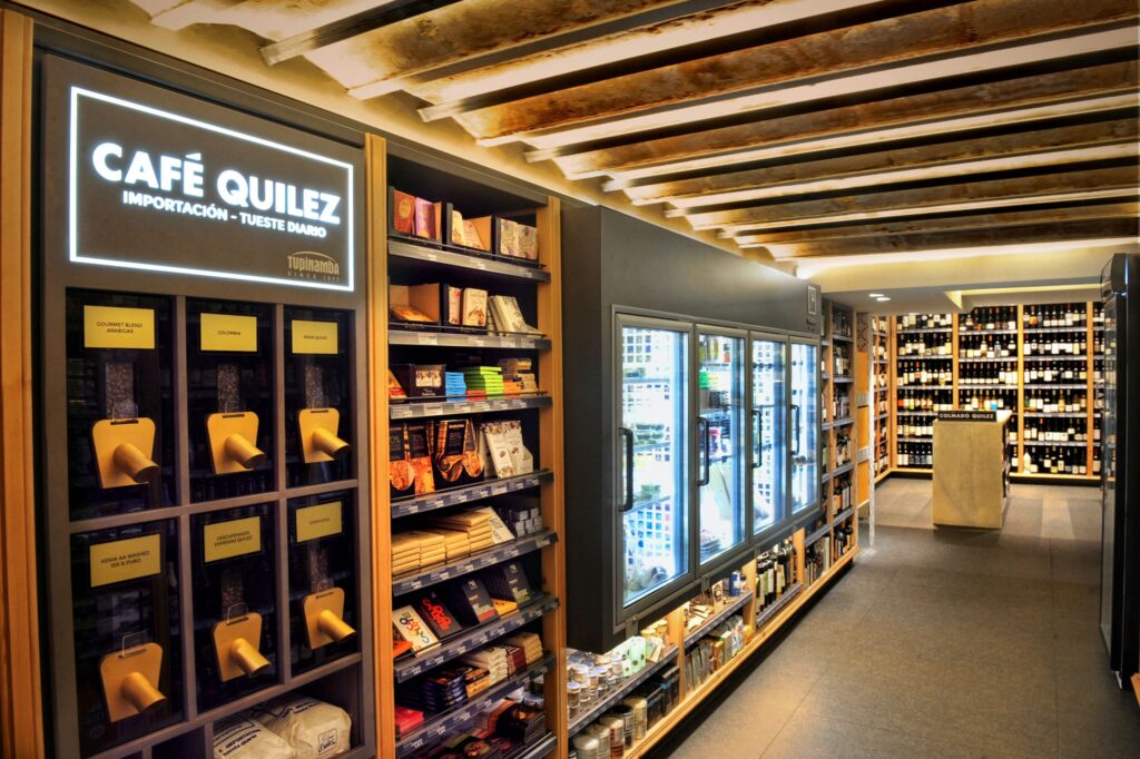 Diseño de interiores de alta gama en Barcelona: Boutique Café Quilez Vista interior de la boutique contemporánea Café Quilez en Barcelona, mostrando un elegante diseño con estanterías de madera y unidades de refrigeración de vidrio, iluminada de manera natural y destacando productos gourmet.