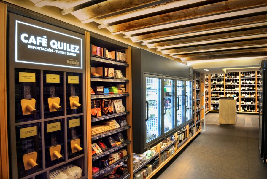 Vista interior de la boutique contemporánea Café Quilez en Barcelona, mostrando un elegante diseño con estanterías de madera y unidades de refrigeración de vidrio, iluminada de manera natural y destacando productos gourmet.