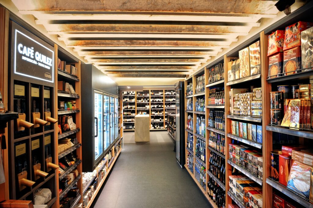 Vista interior de tienda gourmet en Barcelona - Diseño moderno y funcionalidad Vista interior de una tienda de productos gourmet en Barcelona, con estantes llenos de vinos, chocolates y alimentos de alta calidad. Diseño contemporáneo con iluminación suave y techo de vigas expuestas, reflejando el estilo local.