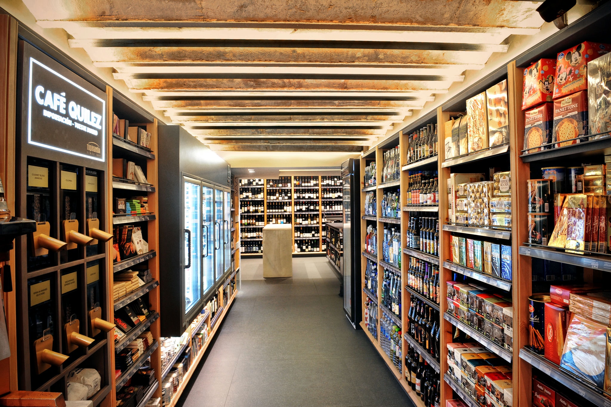 Vista interior de una tienda de productos gourmet en Barcelona, con estantes llenos de vinos, chocolates y alimentos de alta calidad. Diseño contemporáneo con iluminación suave y techo de vigas expuestas, reflejando el estilo local.