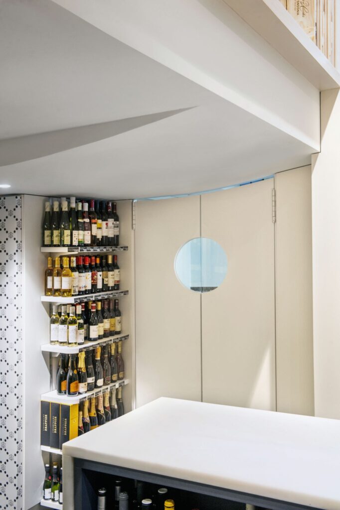 Proyecto de diseño de interiores moderno en Barcelona: área de almacenamiento de vino elegante Interior de un proyecto de diseño de interiores en Barcelona que muestra un área de almacenamiento de vino moderna con estantes organizados. Las paredes tienen texturas sutiles y el techo presenta líneas angulares contemporáneas. Una ventana circular destaca la sofisticación arquitectónica, combinando funcionalidad y estética con materiales de alta calidad como madera y mármol.