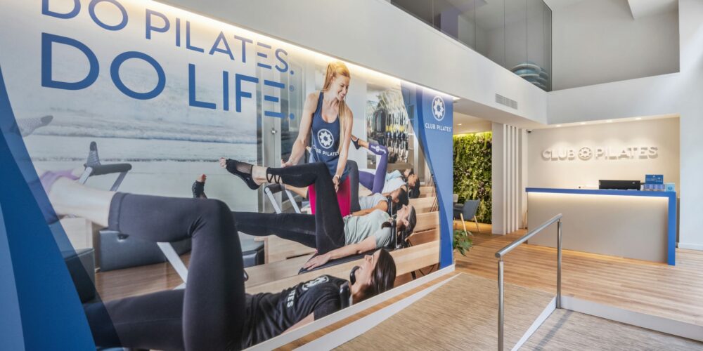 Club Pilates Bonanova: El interior de un moderno estudio de pilates con un área de recepción acogedora, un gran mural con el eslogan '¡HAZ PILATES. ¡HAZ VIDA!' y un instructor de pilates asistiendo a clientes en equipos de reformador.