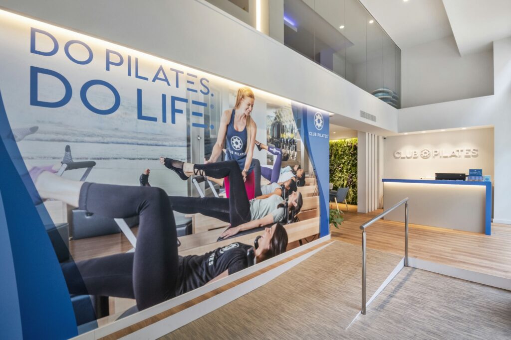 Club Pilates Bonanova: El interior de un moderno estudio de pilates con un área de recepción acogedora, un gran mural con el eslogan '¡HAZ PILATES. ¡HAZ VIDA!' y un instructor de pilates asistiendo a clientes en equipos de reformador.