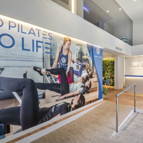 Club Pilates Bonanova: El interior de un moderno estudio de pilates con un área de recepción acogedora, un gran mural con el eslogan '¡HAZ PILATES. ¡HAZ VIDA!' y un instructor de pilates asistiendo a clientes en equipos de reformador.