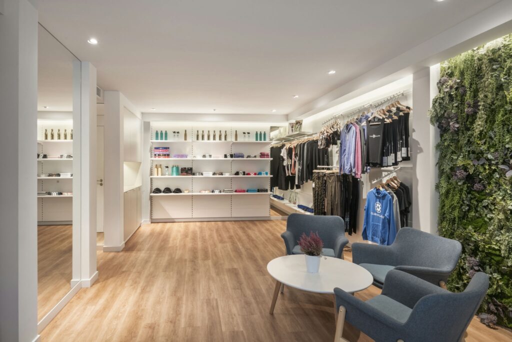 Espacio de venta minorista boutique moderno con atmósfera acogedora y decoración elegante Interior de una tienda minorista moderna con suelos de madera, suave iluminación superior, un gran espejo a la izquierda, una pared vertical de plantas a la derecha y estanterías organizadas con productos de estilo de vida y ropa deportiva. Área de descanso acogedora con dos sillones elegantes alrededor de una mesa pequeña.