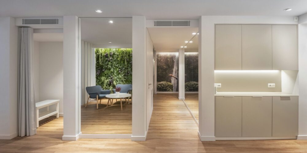 Un interior moderno bellamente diseñado que exhibe un diseño de concepto abierto con transiciones fluidas entre las habitaciones. La imagen presenta un suelo de madera natural, un área de cocina elegante con mobiliario minimalista y una acogedora zona de estar con sillas azules. Paneles de vidrio de piso a techo permiten que entre abundante luz solar, creando una atmósfera brillante y aireada, complementada por una iluminación suave y vegetación.