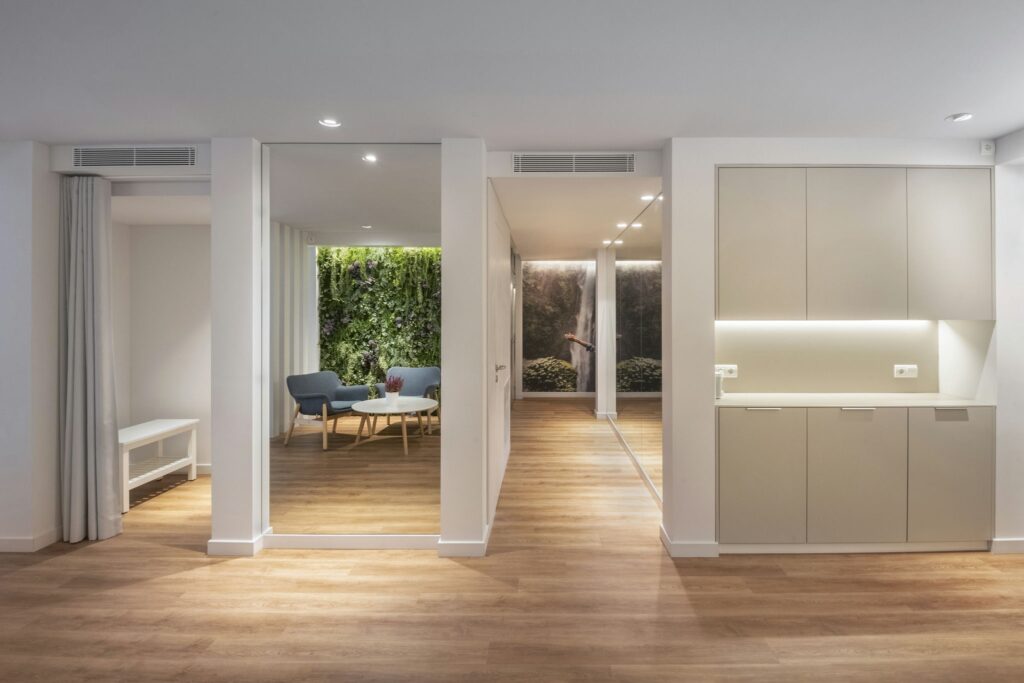 Interior Moderno Abierto y Elegante con Luz Natural Un interior moderno bellamente diseñado que exhibe un diseño de concepto abierto con transiciones fluidas entre las habitaciones. La imagen presenta un suelo de madera natural, un área de cocina elegante con mobiliario minimalista y una acogedora zona de estar con sillas azules. Paneles de vidrio de piso a techo permiten que entre abundante luz solar, creando una atmósfera brillante y aireada, complementada por una iluminación suave y vegetación.