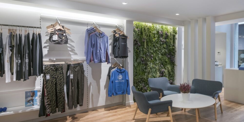 Un espacio minorista moderno que muestra una exhibición bien organizada de ropa deportiva colorida, incluyendo leggings y ropa activa de la marca 'Club Pilates', dispuesta en percheros de madera minimalistas contra una elegante pared blanca. Dos cómodas sillas grises están colocadas prominentemente en el primer plano, acompañadas de una mesa circular blanca y una pequeña planta en maceta, mientras que un jardín vertical con una exuberante vegetación proporciona un vibrante fondo complementado por un cálido suelo de madera.