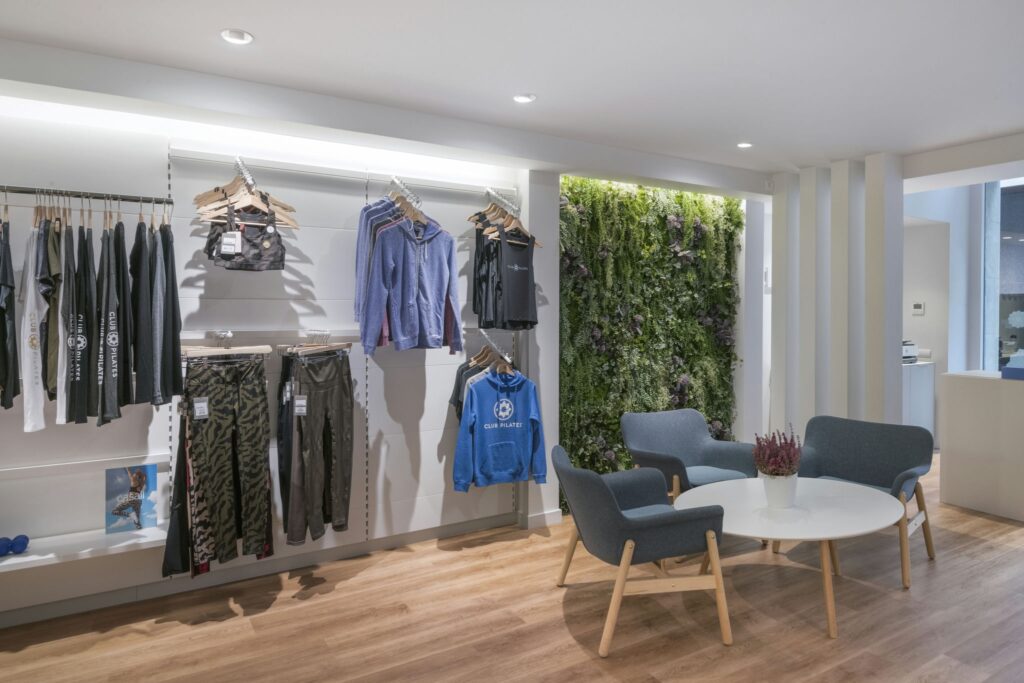 Exhibición de Ropa Deportiva Estilizada en un Espacio Minorista Moderno Un espacio minorista moderno que muestra una exhibición bien organizada de ropa deportiva colorida, incluyendo leggings y ropa activa de la marca 'Club Pilates', dispuesta en percheros de madera minimalistas contra una elegante pared blanca. Dos cómodas sillas grises están colocadas prominentemente en el primer plano, acompañadas de una mesa circular blanca y una pequeña planta en maceta, mientras que un jardín vertical con una exuberante vegetación proporciona un vibrante fondo complementado por un cálido suelo de madera.