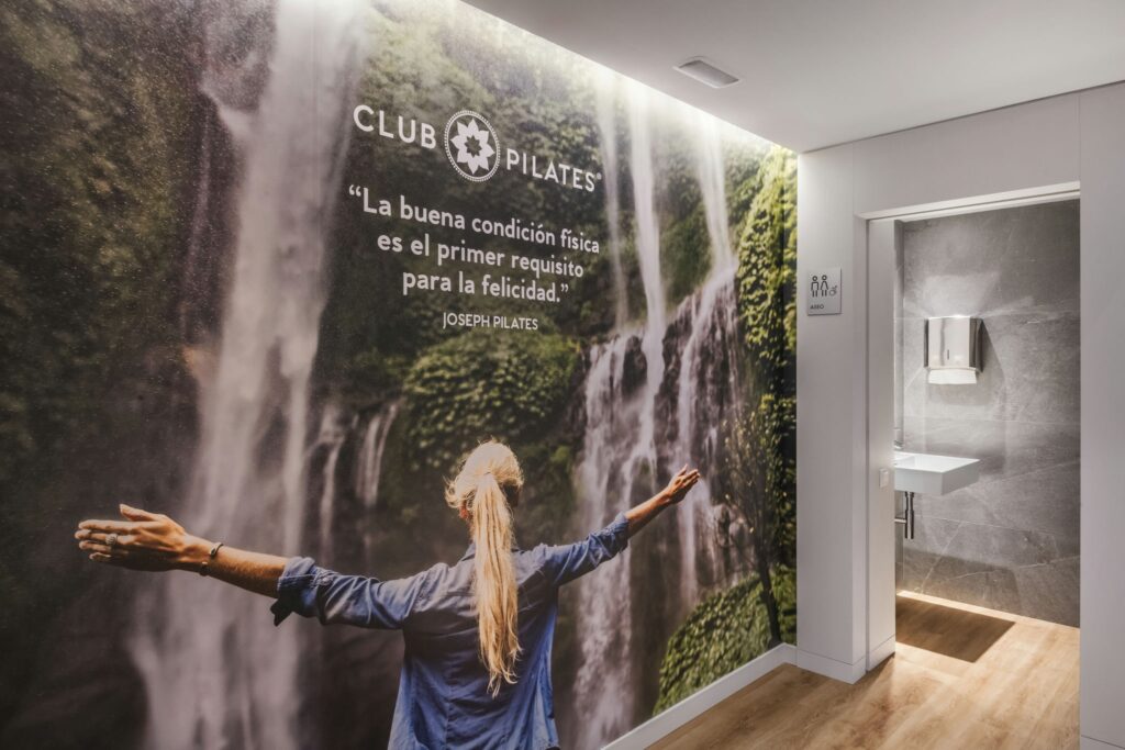 Espacio Interior Tranquilo con Mural de Cascada y Cita Motivacional | Estudio de Bienestar Un sereno estudio de bienestar interior que presenta un vibrante mural que representa una cascada en caída rodeada de exuberante vegetación, simbolizando la tranquilidad. Una persona con cabello largo, vestida con una camisa azul, se encuentra con los brazos extendidos, abrazando la sensación de libertad y conexión con la naturaleza. Una cita motivacional de Joseph Pilates en español enfatiza el bienestar físico para la felicidad. Un área de baño limpia y moderna es visible en el fondo, mejorando la atmósfera acogedora.