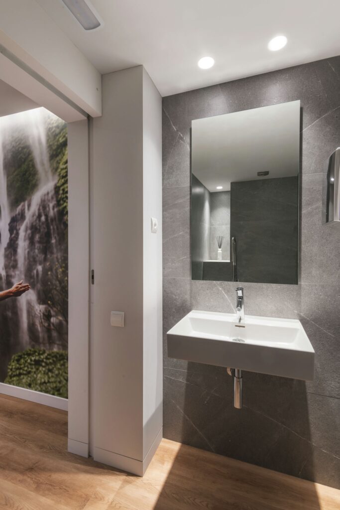 Baño moderno con lavabo montado en la pared y fondo de cascada escénica Un baño contemporáneo que presenta un elegante lavabo montado en la pared con un grifo estiloso, azulejos de pared gris oscuro, un gran espejo que refleja una iluminación suave y un singular fondo de cascada a un lado, complementado por un suelo de madera clara.