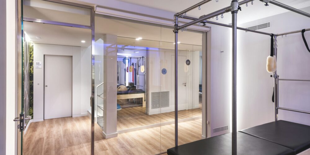 Un estudio de Pilates luminoso y contemporáneo que cuenta con un reformador de Pilates en primer plano, paredes de vidrio, suelo de madera y una pared espejo que muestra el equipo de entrenamiento. El espacio está diseñado para actividades de fitness y bienestar, complementado con una iluminación ambiental suave y relajante.