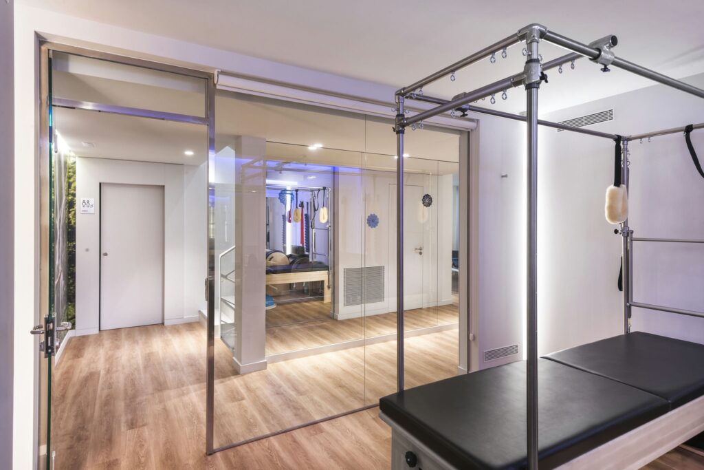 Estudio de Pilates Moderno con Reformas y Iluminación Ambiental Un estudio de Pilates luminoso y contemporáneo que cuenta con un reformador de Pilates en primer plano, paredes de vidrio, suelo de madera y una pared espejo que muestra el equipo de entrenamiento. El espacio está diseñado para actividades de fitness y bienestar, complementado con una iluminación ambiental suave y relajante.