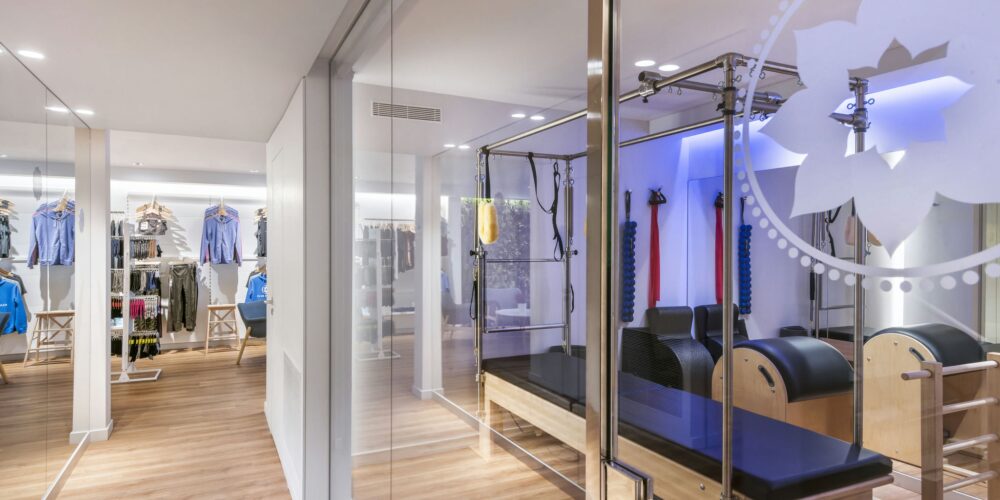 Imagen de un moderno estudio de pilates en Barcelona con paredes de vidrio, iluminado naturalmente. Equipos de pilates de alta gama y estanterías de ropa deportiva resaltan la funcionalidad y estética del diseño interior.