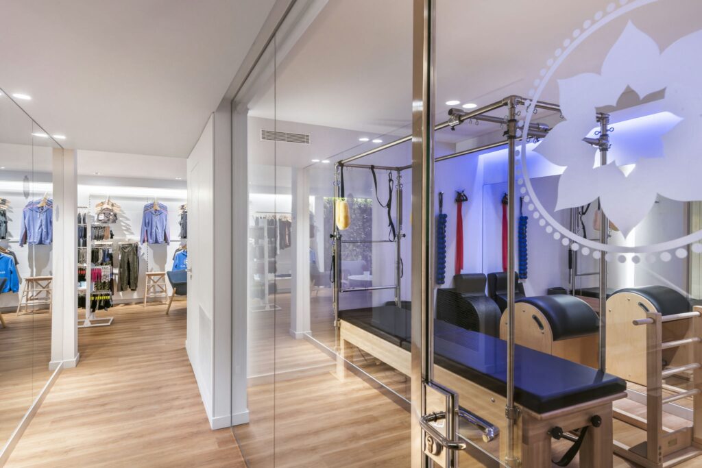 Estudio de Pilates Moderno en Barcelona - Diseño Interior de Alta Gama Imagen de un moderno estudio de pilates en Barcelona con paredes de vidrio, iluminado naturalmente. Equipos de pilates de alta gama y estanterías de ropa deportiva resaltan la funcionalidad y estética del diseño interior.