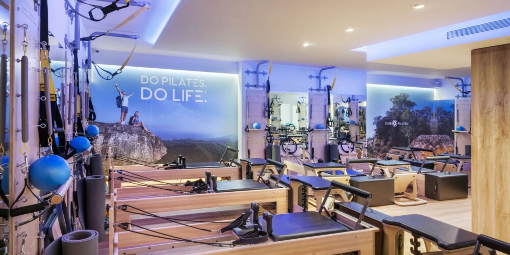 Vista interior de un estudio de Pilates moderno en Barcelona, con suelos de madera, equipo de Pilates de calidad y una atmósfera contemporánea. Las paredes muestran imágenes motivacionales y la frase 'DO PILATES. DO LIFE.', destacando un entorno profesional e invitador para entusiastas del fitness.