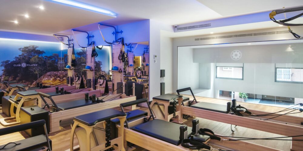 Interior de un estudio de Pilates moderno en Barcelona, con máquinas reformer de madera de alta calidad, iluminación azul suave y espejo grande que refleja arte mural vibrante de paisaje natural.