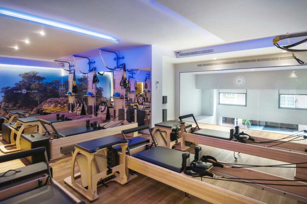 Estudio de Pilates Moderno en Barcelona con Reformers de Madera Interior de un estudio de Pilates moderno en Barcelona, con máquinas reformer de madera de alta calidad, iluminación azul suave y espejo grande que refleja arte mural vibrante de paisaje natural.