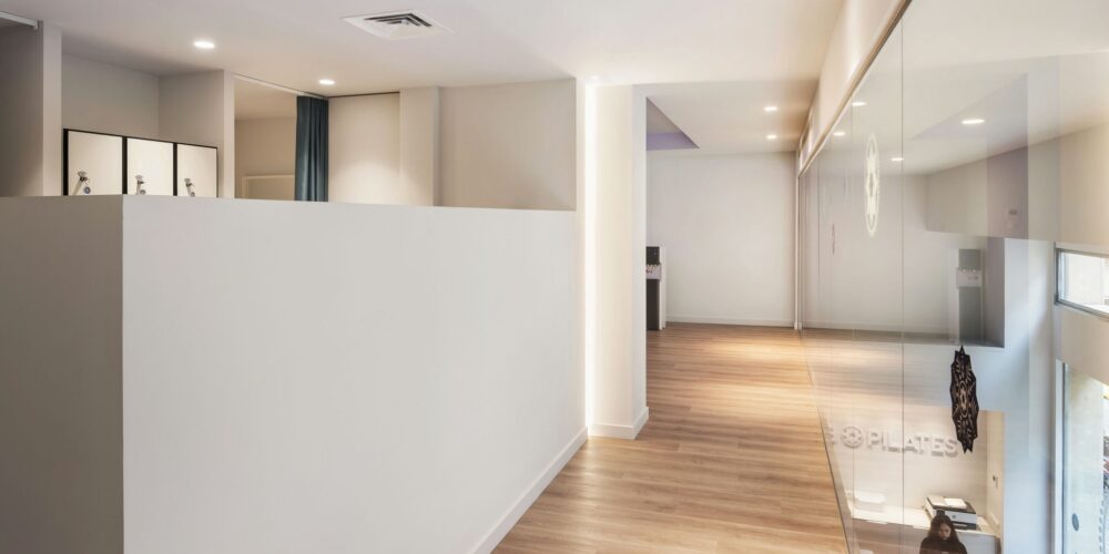 Moderno pasillo de diseño interior en Barcelona con suelos de madera pulida, paredes blancas y grandes paneles de cristal que permiten la entrada de luz natural.
