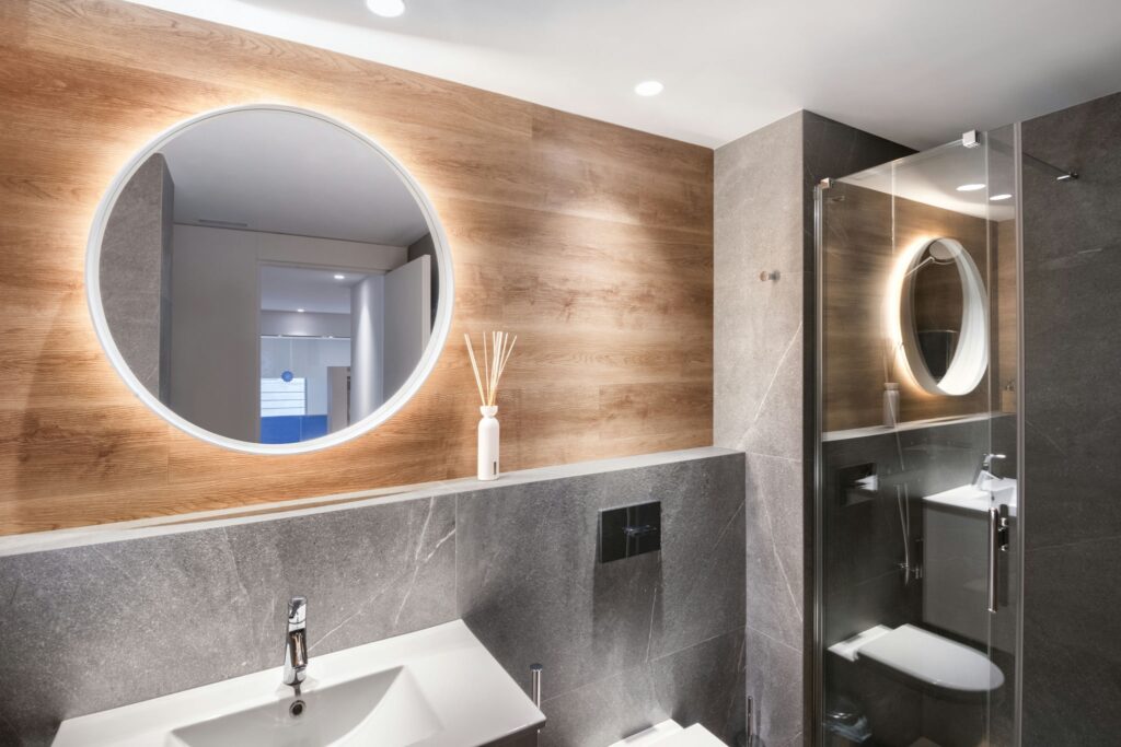 Baño moderno de alta gama en Barcelona con diseño elegante Interior de baño contemporáneo en Barcelona, con espejo circular retroiluminado, pared de madera y lavabo elegante de mármol gris.